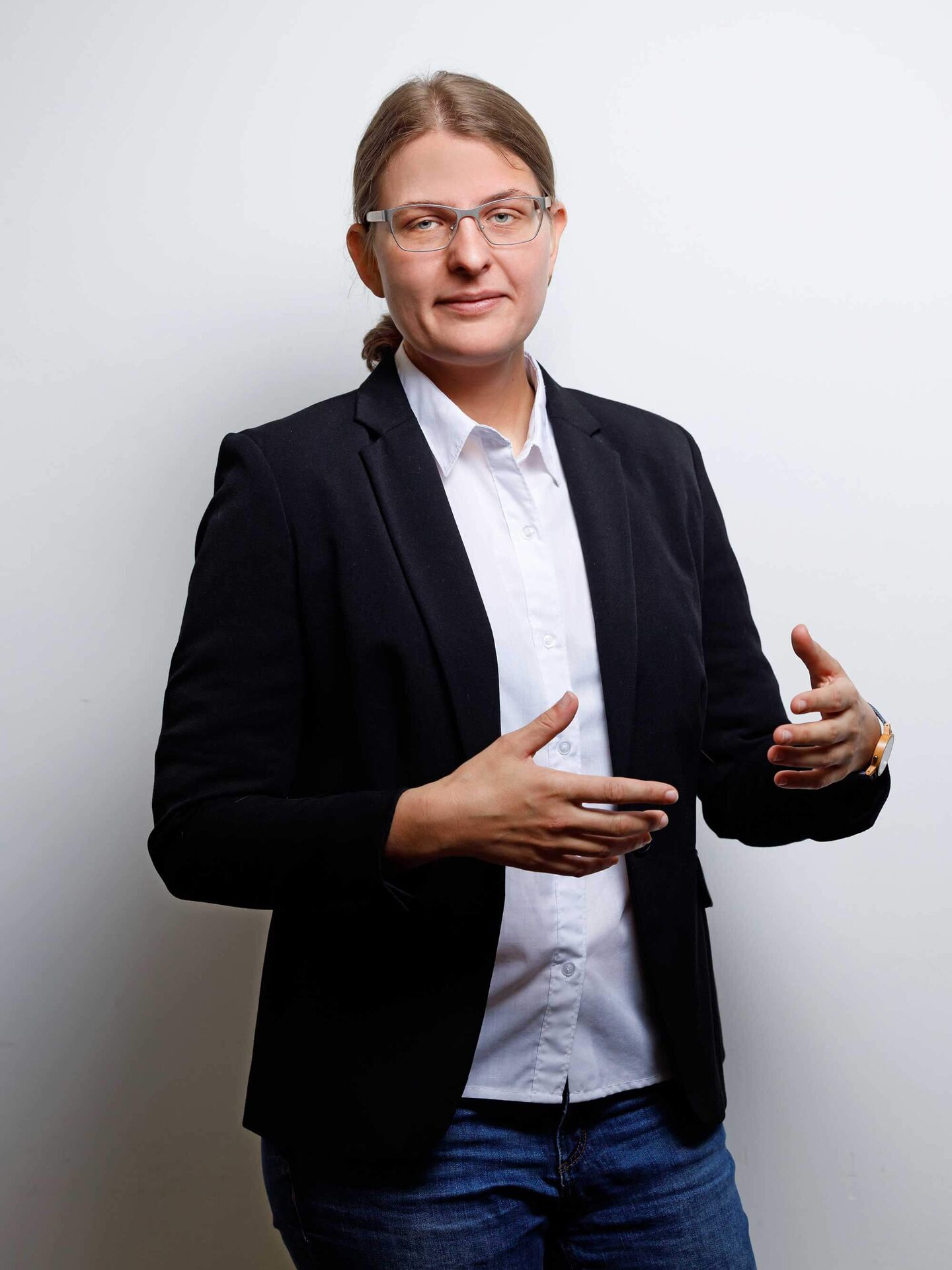 Laureen ist mit einer weißen Bluse, einem schwarzen Blazer und einer blauen Jeans bekleidet. Sie gestikuliert und schaut in die Kamera.
