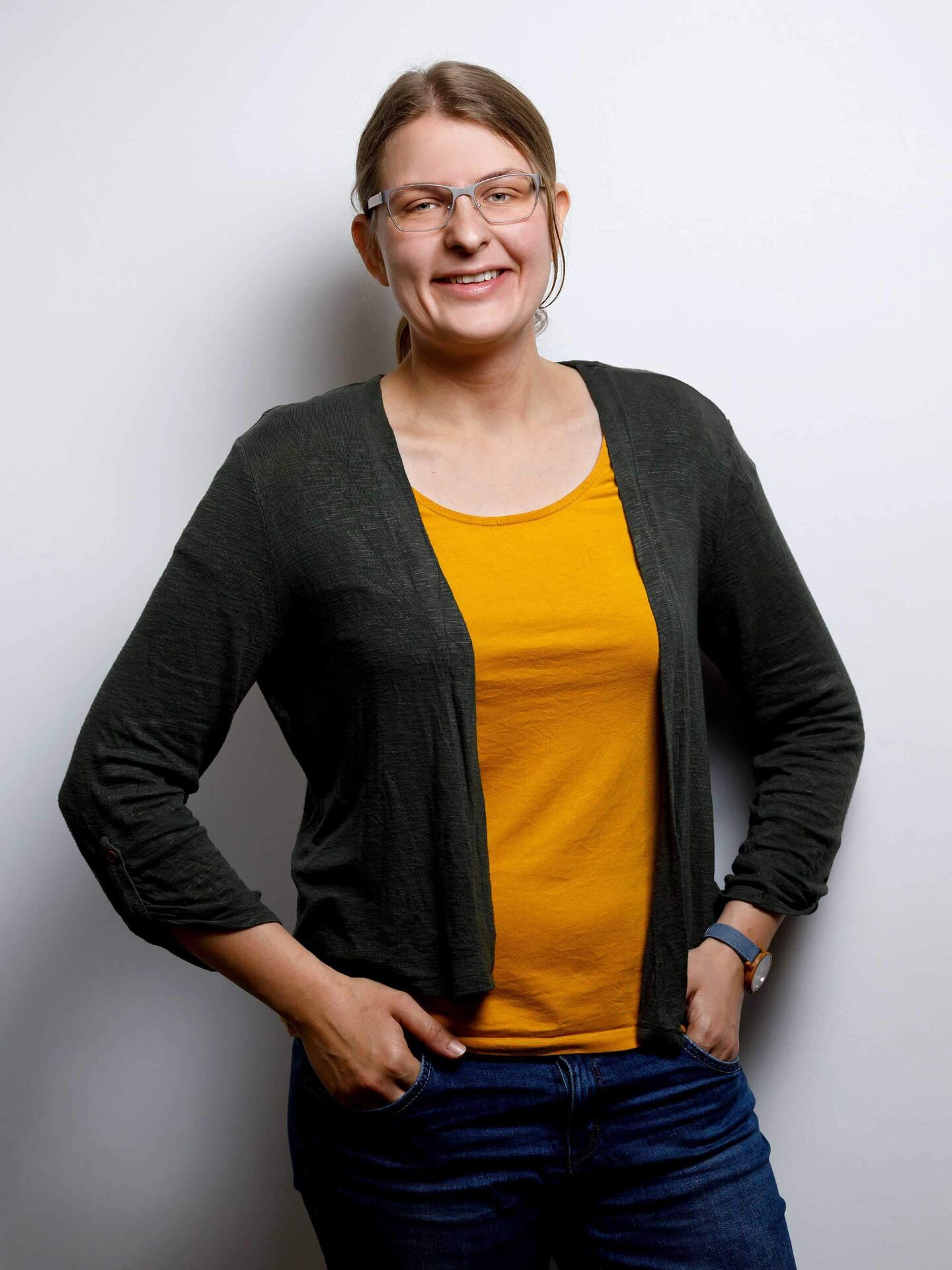 Laureen ist mit einem geleben T-Shirt, einer dunkelgrünen Strickjacke und einer blauen Jeans bekleidet. Sie hat die Hände in den vorderen Hosentaschen und lächelt in die Kamera.