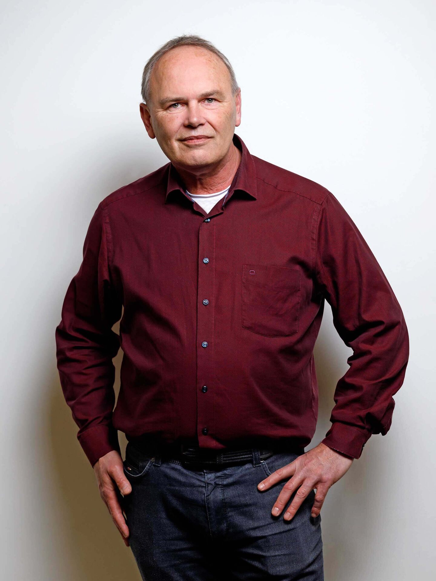 Sven ist mit einem roten Hemd und einer dunklen Jeans bekleidet. Am Halsausschnitt ist ein weißes T-Shirt zu sehen. Er hat seine Hände an die Hosentaschen gelegt.