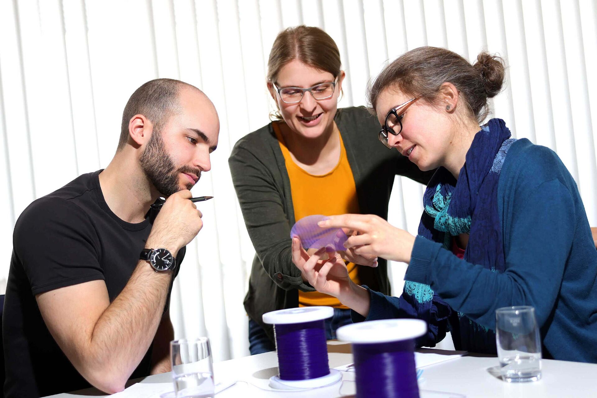 Three people sit and stand around a table with water glasses, pens and pads, and spools of violet material. One staff member holds a violet mesh in her hand and shows it to the other two people.