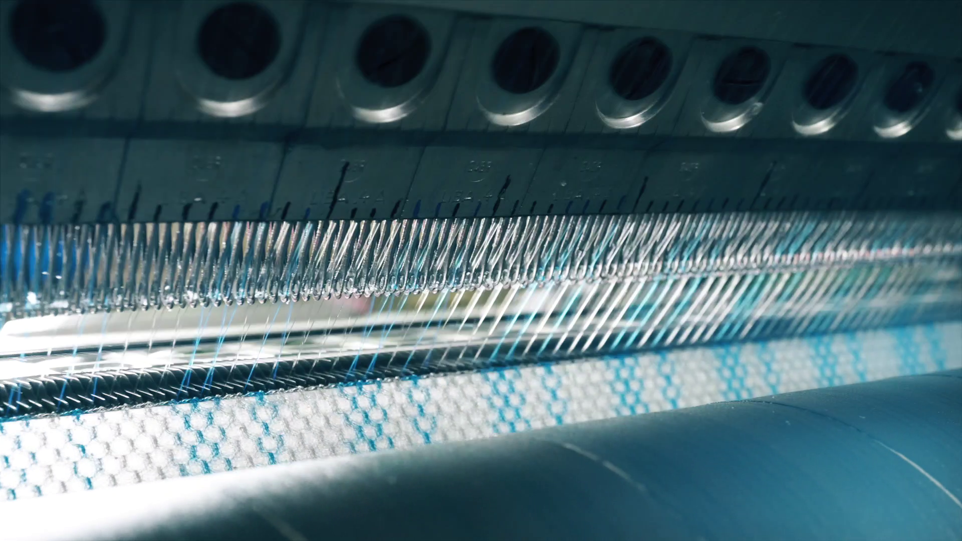 Es ist ein Ausschnitt der Netzherstellung an der Wirkmaschine abgebildet. im Unteren Bereich ist ein Teil des Netzes zu sehen, Oberhalb davon die einzelnen Filamente, die durch Nadeln laufen.