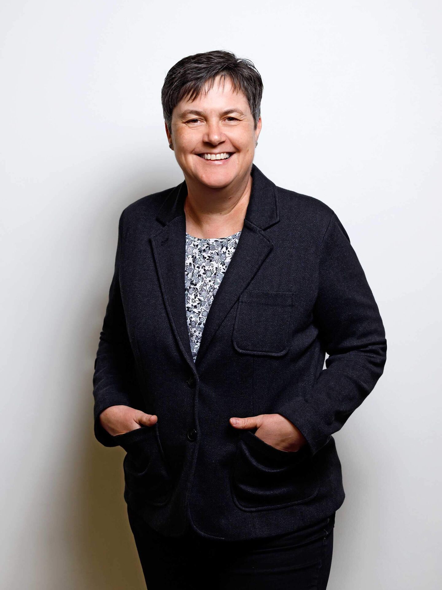 Kathrin trägt ein gemustertes Oberteil zu einem schwarzen Blazer und einer schwarzen Hose. Sie hat die Hände in den Blazertaschen und lächelt in die Kamera.