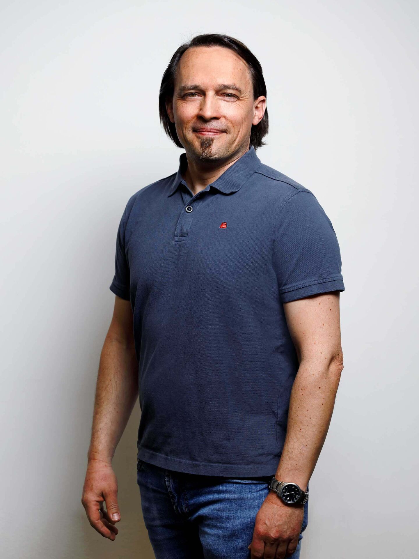 Tobias trägt ein dunkelblaues Poloshirt und eine blaue Jeans. Seine Hände hängen herunter. Er lächelt in die Kamera.