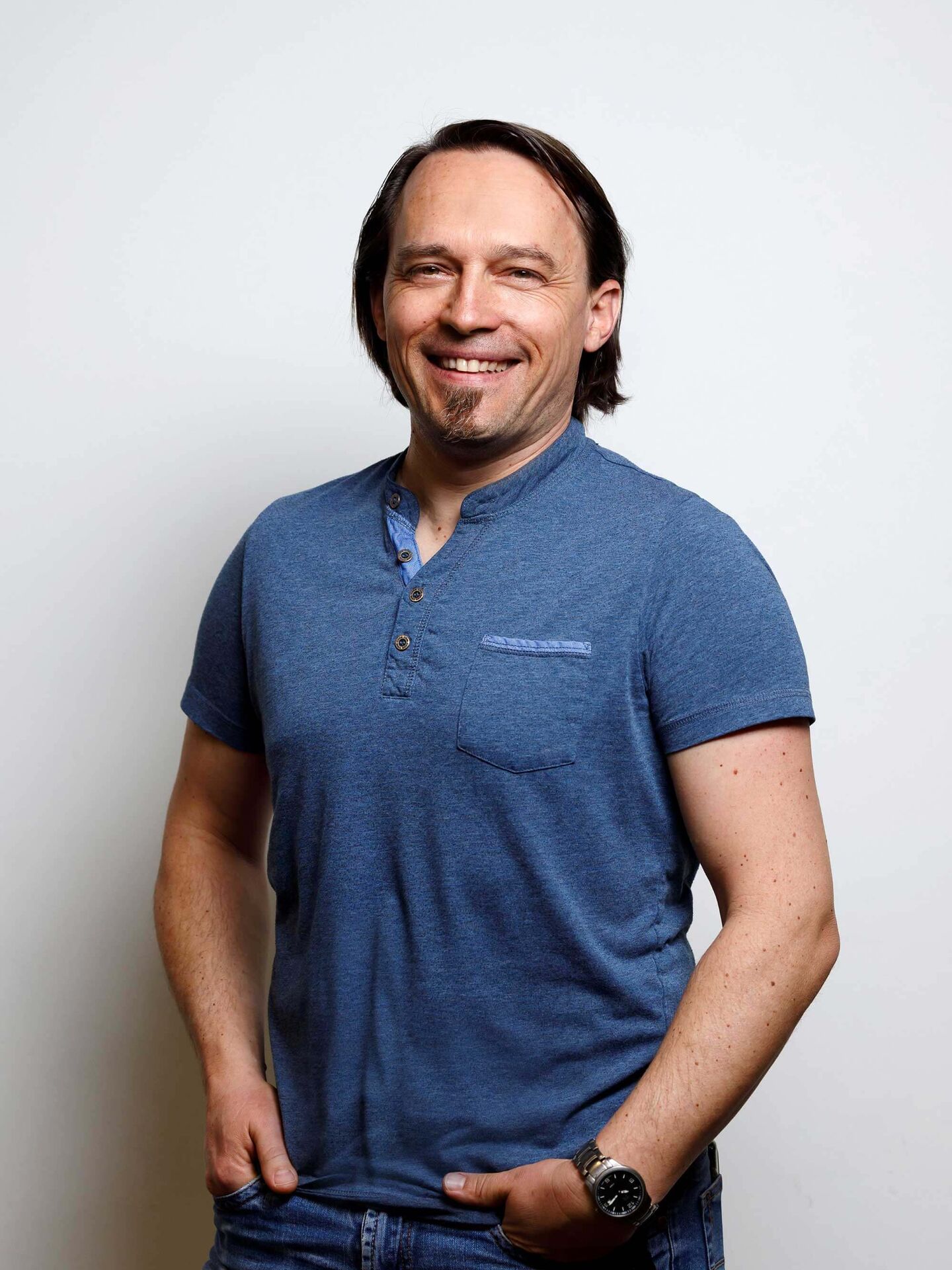 Tobias trägt ein blaues T-Shirt und eine blaue Jeans. Seine Hände sind in den vorderen Hosentaschen. Er lacht in die Kamera.