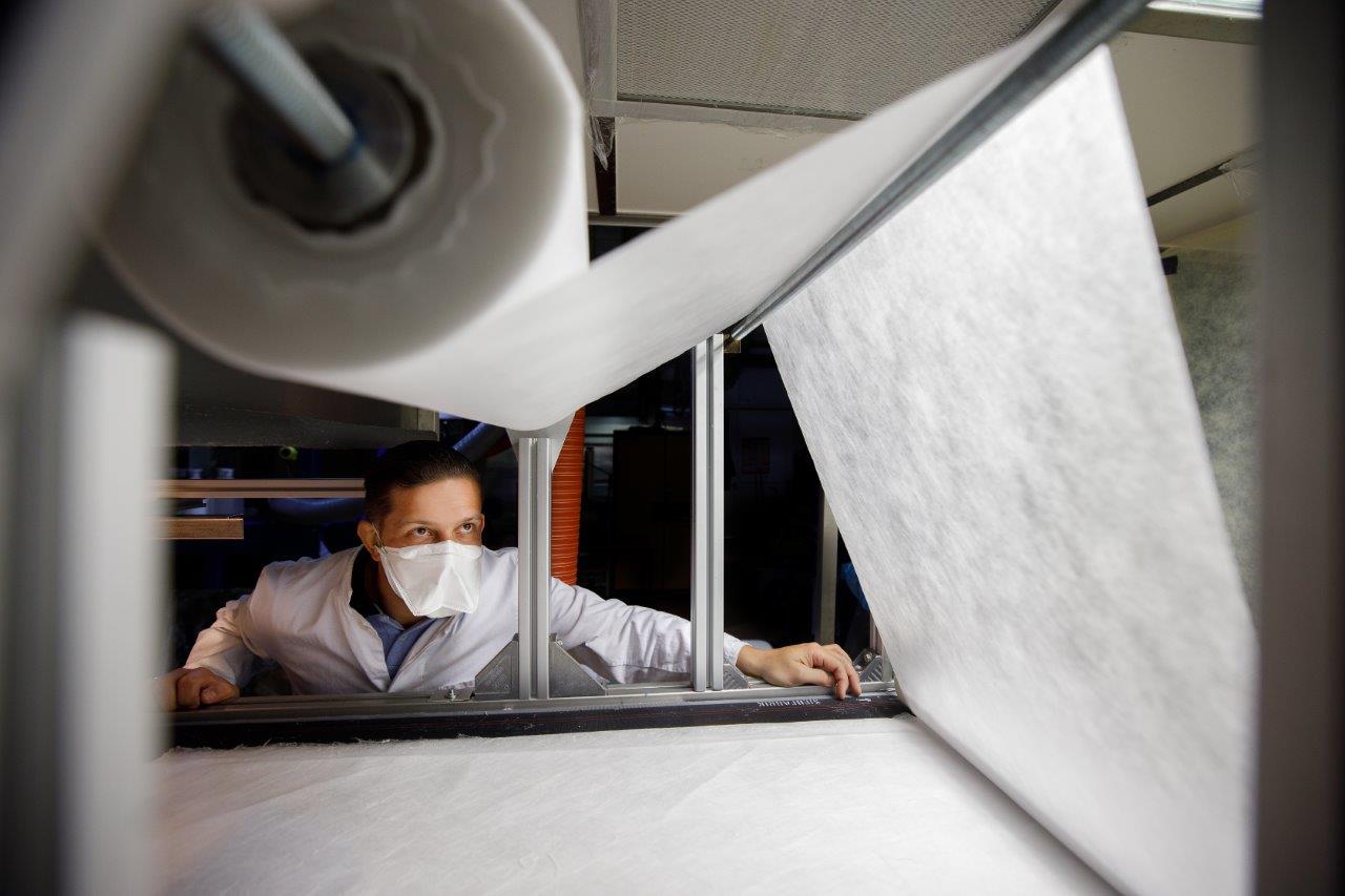 An employee wearing a face mask and white coat looks up. At the top left is a roll of nonwoven, which is unwound over bars from the top right to the bottom right. At the bottom is another layer of nonwoven.